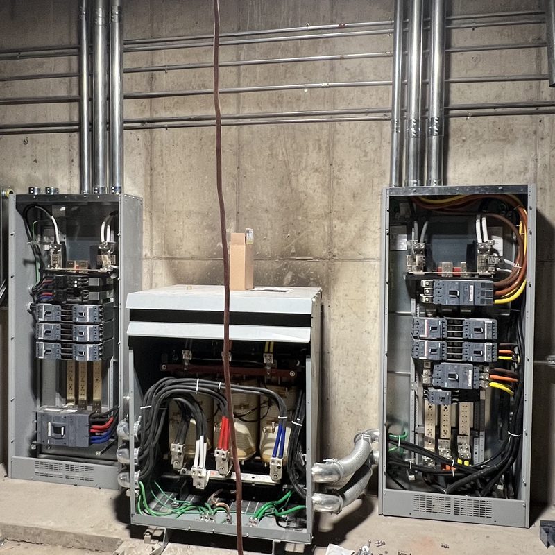 Two open switchboard sections either side of a transformer, with conduit racks running up a concrete wall.